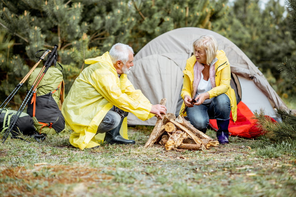 Senior couple in yellow raincoats making fireplace at the campsite near the tent in the woods
