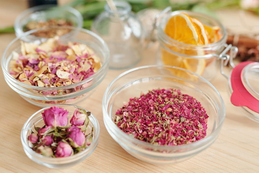 Three bowls with dry rose petals and rosebuds and small jar with orange slices near by on wooden table