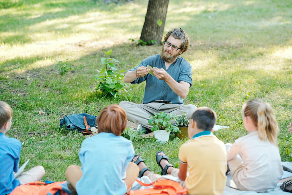 Young botany teacher in glasses holding plant leaf and explaining photosynthesis process to pupils at outdoor class