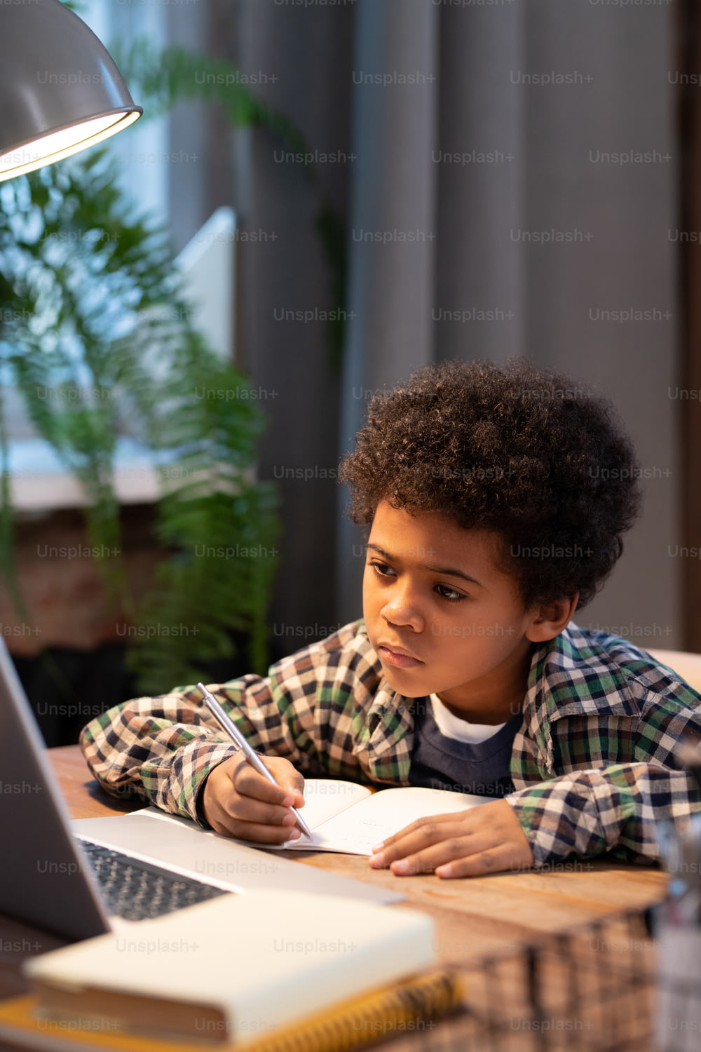 Colegial africano serio que toma notas en el cuaderno mientras está sentado junto a la mesa frente a la computadora portátil y mira la pantalla durante la lección en línea