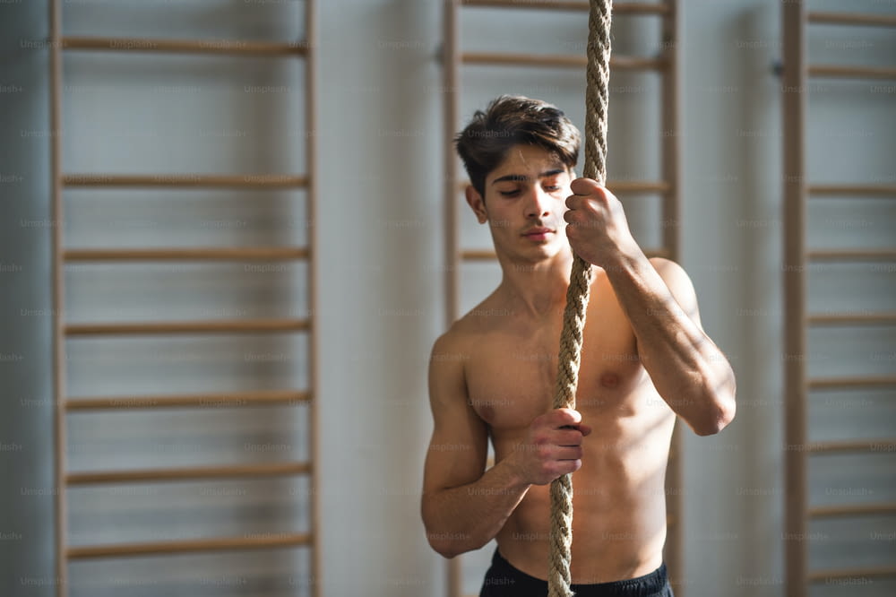 A fit young man in gym standing topless , holding a climbing rope. Copy space.