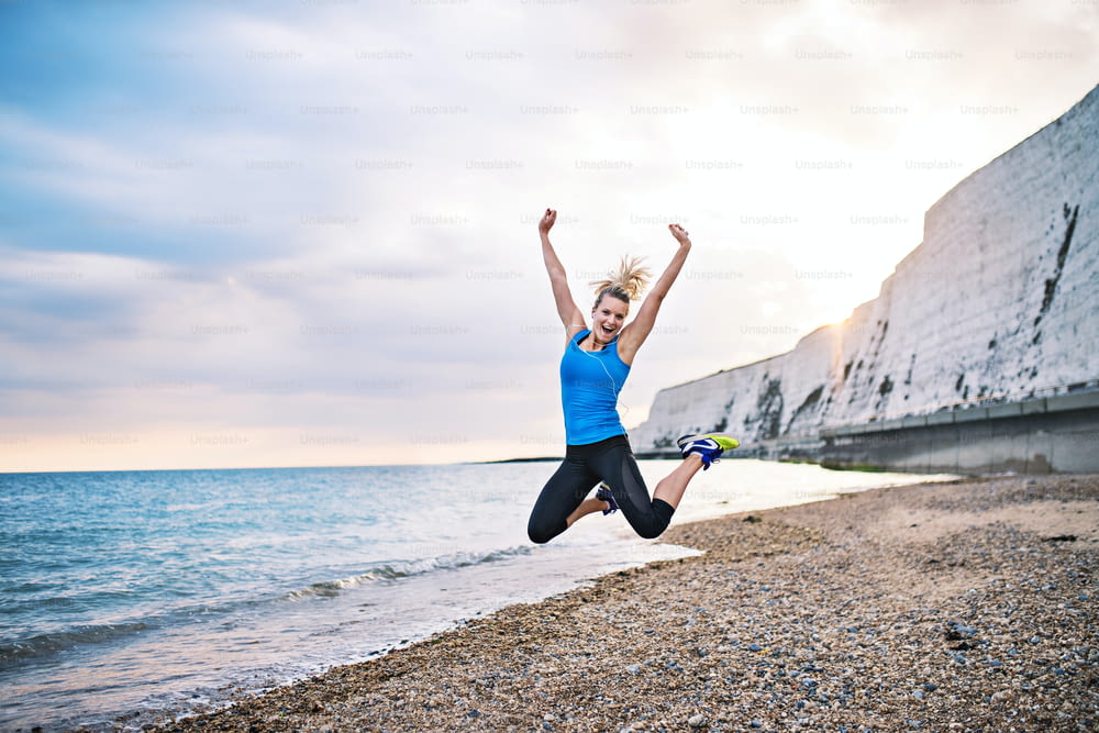 Young sporty woman runner in blue sportswear jumping outside on the beach in nature. Copy space.