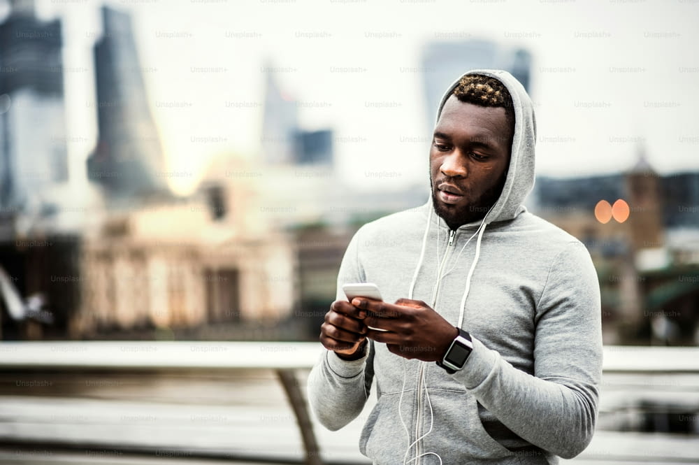 Jovem atleta corredor negro com smartwatch, fones de ouvido e smartphone na ponte em uma cidade, descansando. Espaço de cópia.
