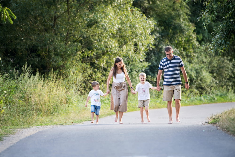 A young family with two small cheerful sons walking barefoot on a road in park on a summer day, holding hands.
