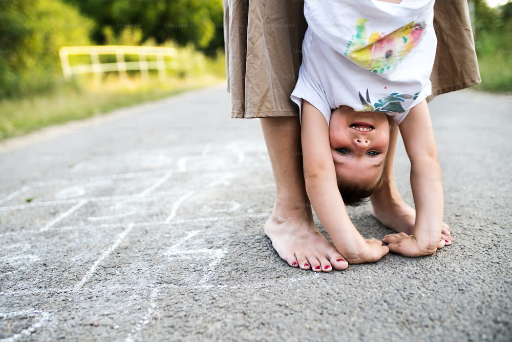 Unrecognizable barefoot mother holding a small son upside down on a road in park on a summer day.
