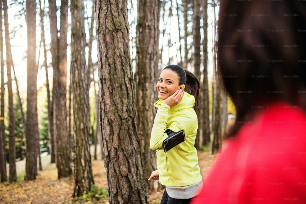Two female runners with earphones and smartphones in armband jogging outdoors in forest in autumn nature, talking.