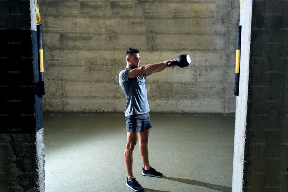 Dedicated muscular man in sportswear doing exercises with kettlebell while standing in gym gym.