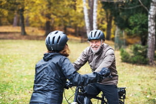 An active senior couple with helmets and electrobikes standing outdoors on a road in nature, looking at each other.