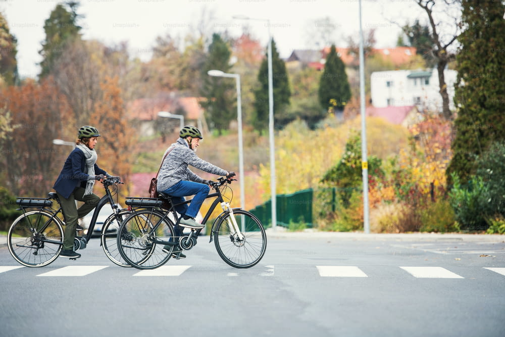 Pareja de ancianos activos con bicicletas eléctricas al aire libre cruzando una calle en la ciudad. Espacio de copia.