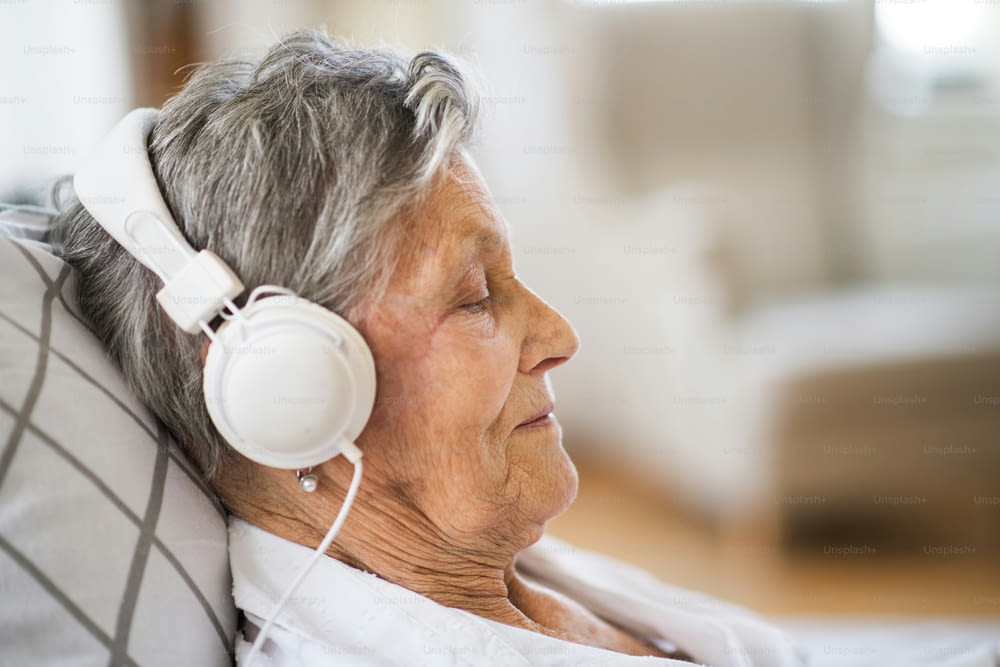 A close-up of sick senior woman with headphones lying in bed at home or in hospital, listening to music.