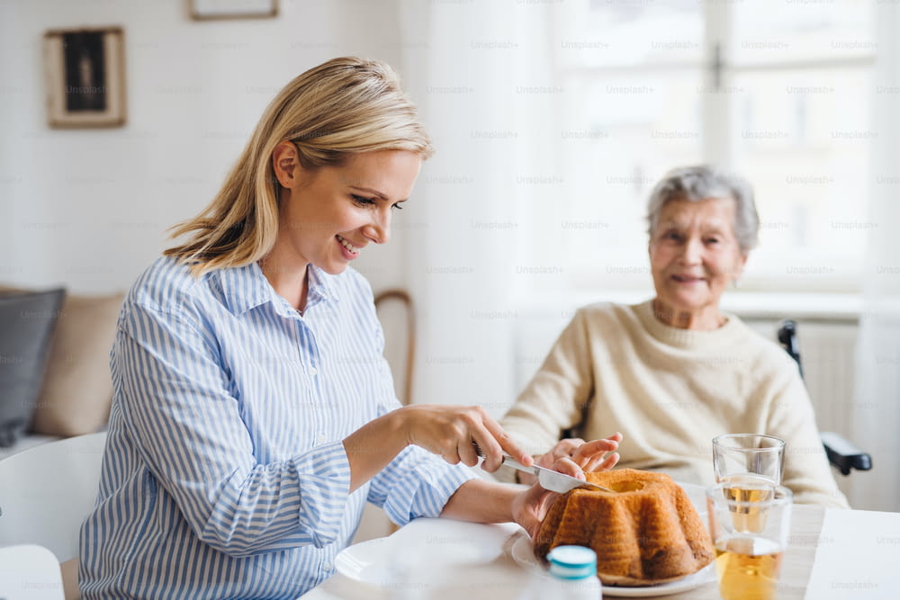 Une femme âgée en fauteuil roulant avec un visiteur de santé assis à la table à la maison, coupant un gâteau.
