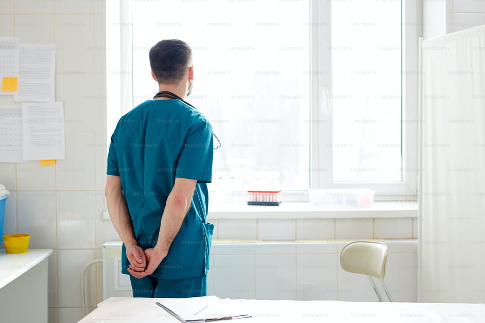 Back view of contemporary clinician in uniform looking through window in hospital