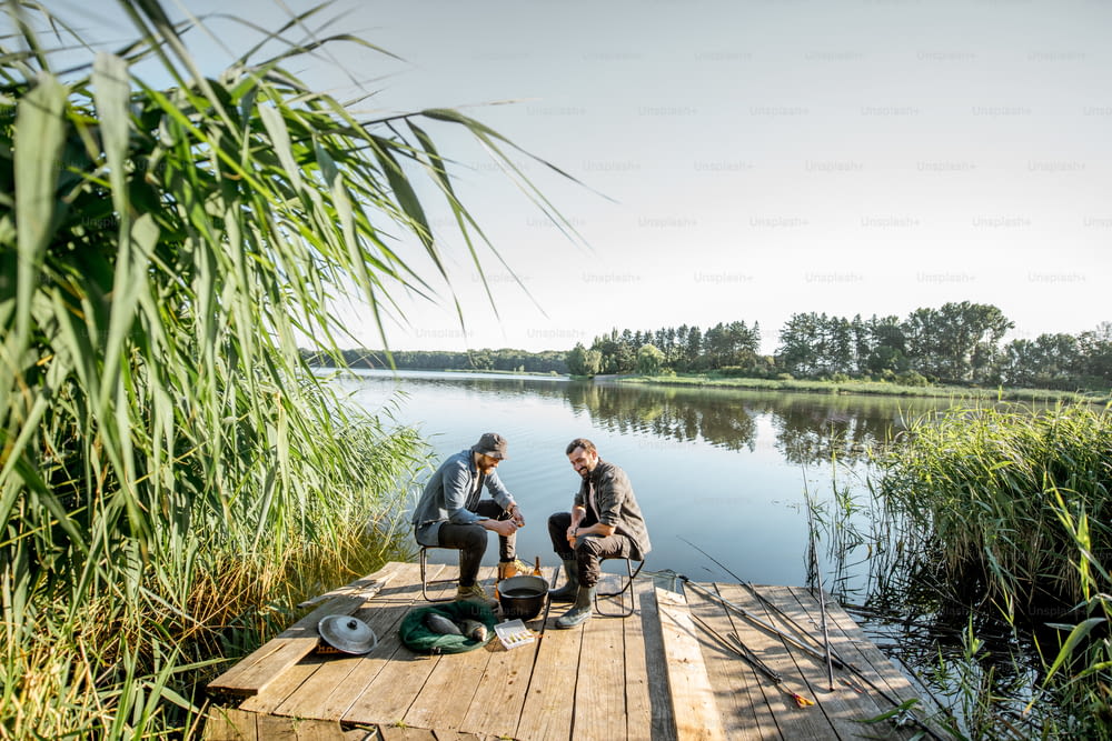 Dos pescadores relajándose durante el picnic en el hermoso muelle de madera con caña verde en el lago por la mañana