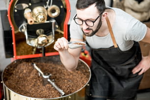 Uomo che controlla la qualità dei chicchi di caffè in piedi con la paletta vicino alla macchina per la tostatura alla torrefazione