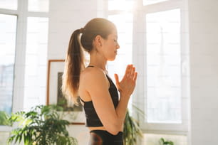 Junge fitte Frau praktiziert Yoga mit Asana Namaste im leichten Yogastudio mit grüner Zimmerpflanze