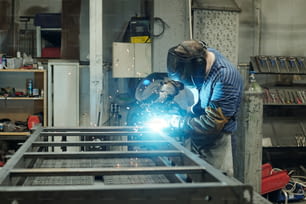 Side view of young male welder in protective face mask carrying out repair or construction work while bending over metallic workbench