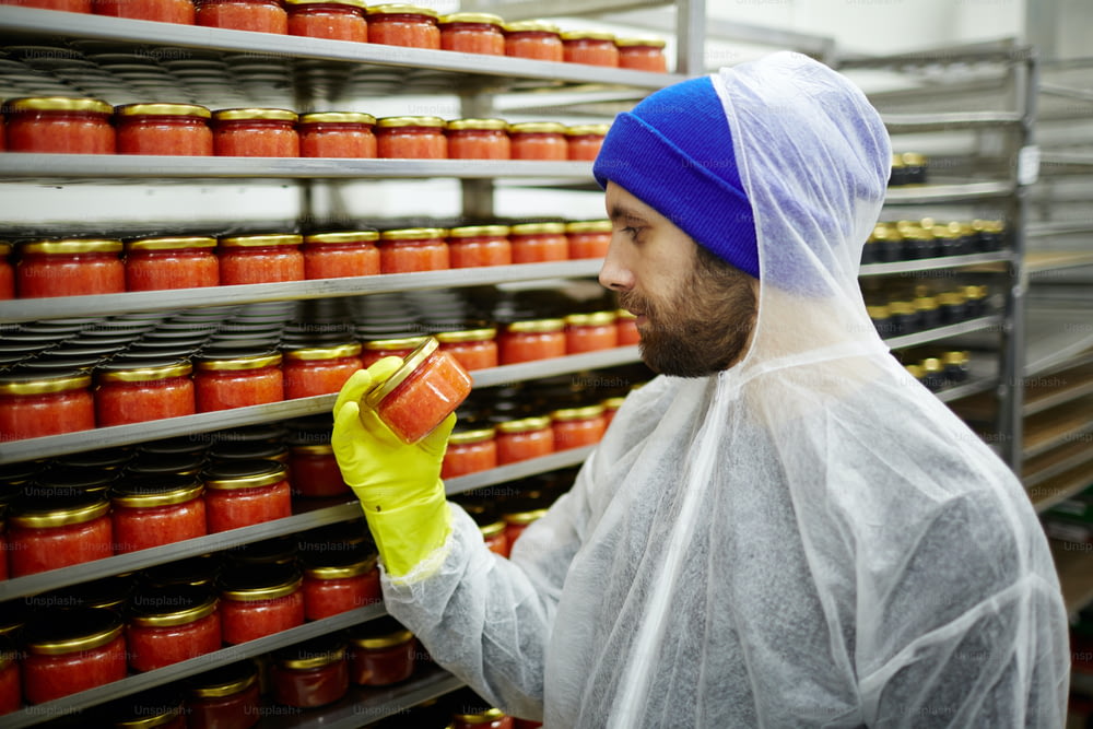Young man in gloves and protective coveralls examining jar with red caviar after packing