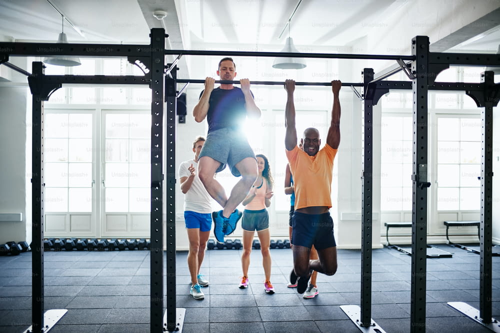 Two fit young men having a friendly pullups competition in a gym with a diverse group of friends cheering in the background