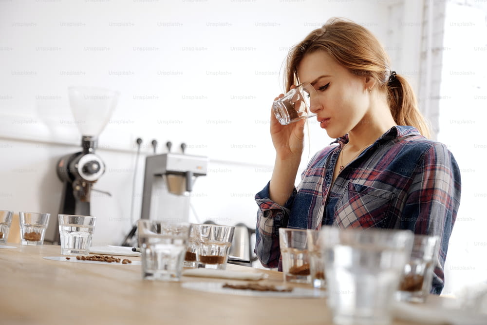 Portrait dans une mise au point sélective d’une jeune barista examinant des tasses avec des grains et du café moulu, faisant un test de dégustation, sentant le café frais