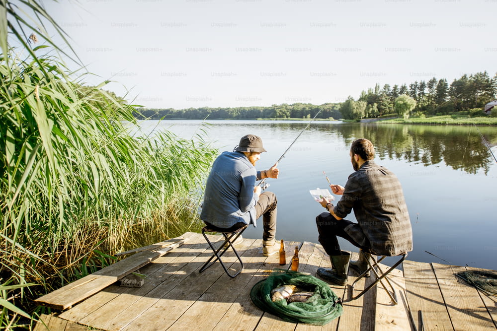 Landschaftsansicht auf den schönen See und grünes Schilf mit zwei Männern, die im Morgenlicht auf dem hölzernen Pier fischen