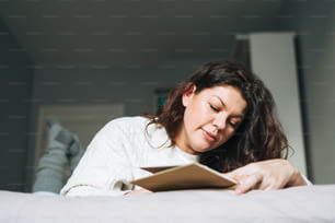 Jeune femme brune adulte de quarante ans plus le corps positif dans un pull tricoté blanc écrit des notes dans le planificateur sur le lit à la maison