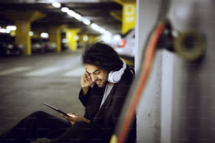 Close up of stylish playful smiling dreadlock skater man sitting leaned on the wall of the underground parking garage in a suit and listening music while holding a tablet.