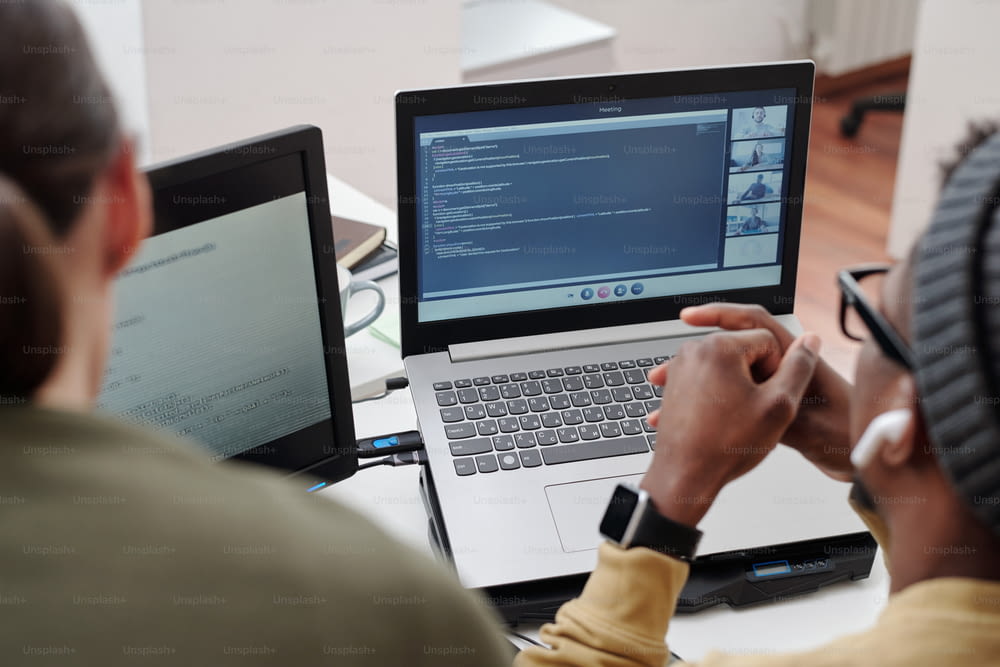 Manos de un joven negro sobre el teclado de la computadora portátil con chat de video en la pantalla mientras está sentado en el lugar de trabajo junto a una colega femenina