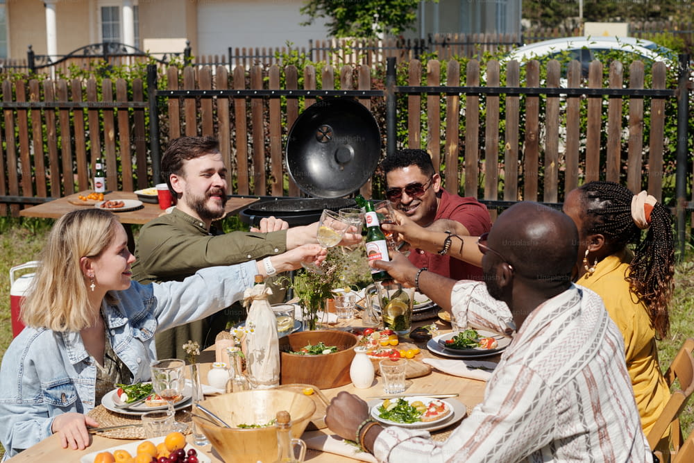 Gruppe junger interrassischer Freunde mit Getränken, die während des Abendessens im Freien oder der Party über dem Tisch mit hausgemachten Speisen und Getränken anstoßen