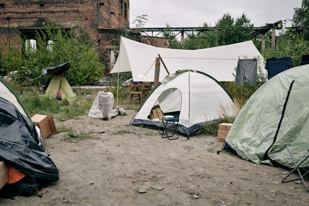Camp de réfugiés avec des tentes, des vêtements suspendus à des cordes et des chaises pliantes contre un bâtiment abandonné
