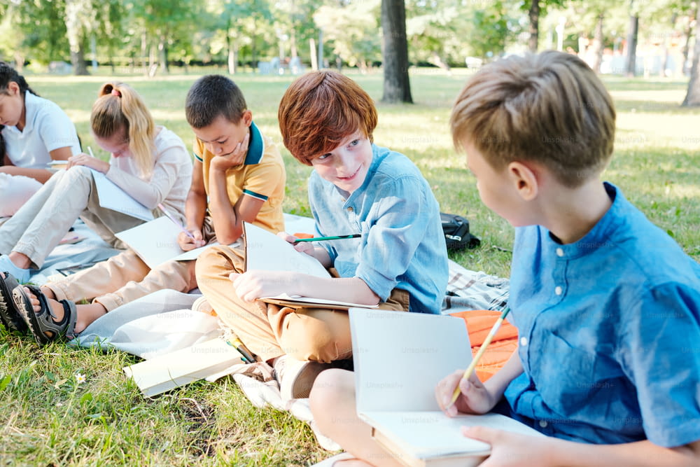 Redhead boy sitting in row of classmates and showing test answers to friend at outdoor lesson, cheating concept