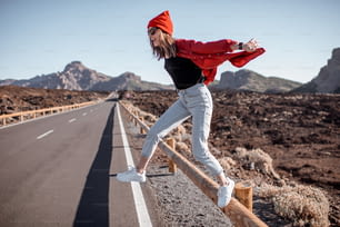Lifestyle portrait of a young woman stylishly dressed in red jumping on the road, traveling on the volcanic valley on a sunny day. Carefree lifestyle and travel concept