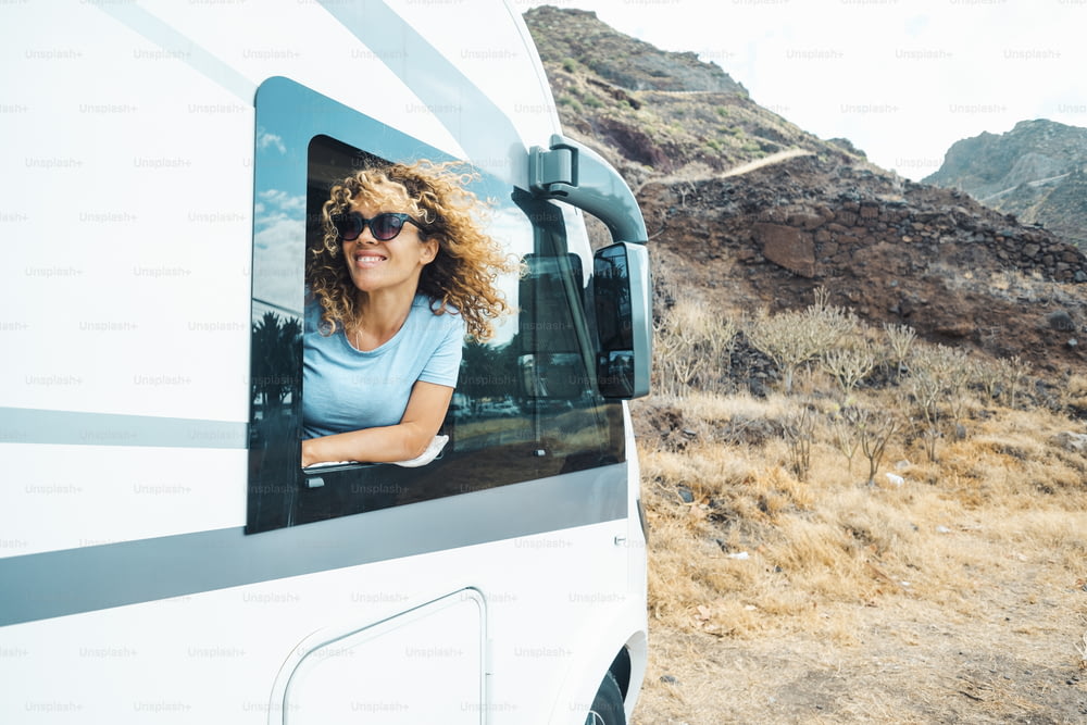 People and travel holiday vacation destination. Happy young adult woman smile and enjoy nature off road parking outside the window of her modern motor home camper van. Female traveler