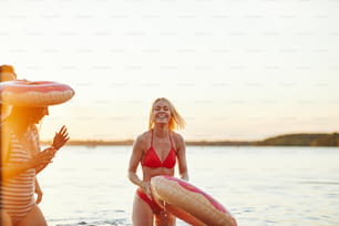 Laughing young blonde woman in a bikini standing in a lake holding a swim ring while having fun with friends at a lake at sunset