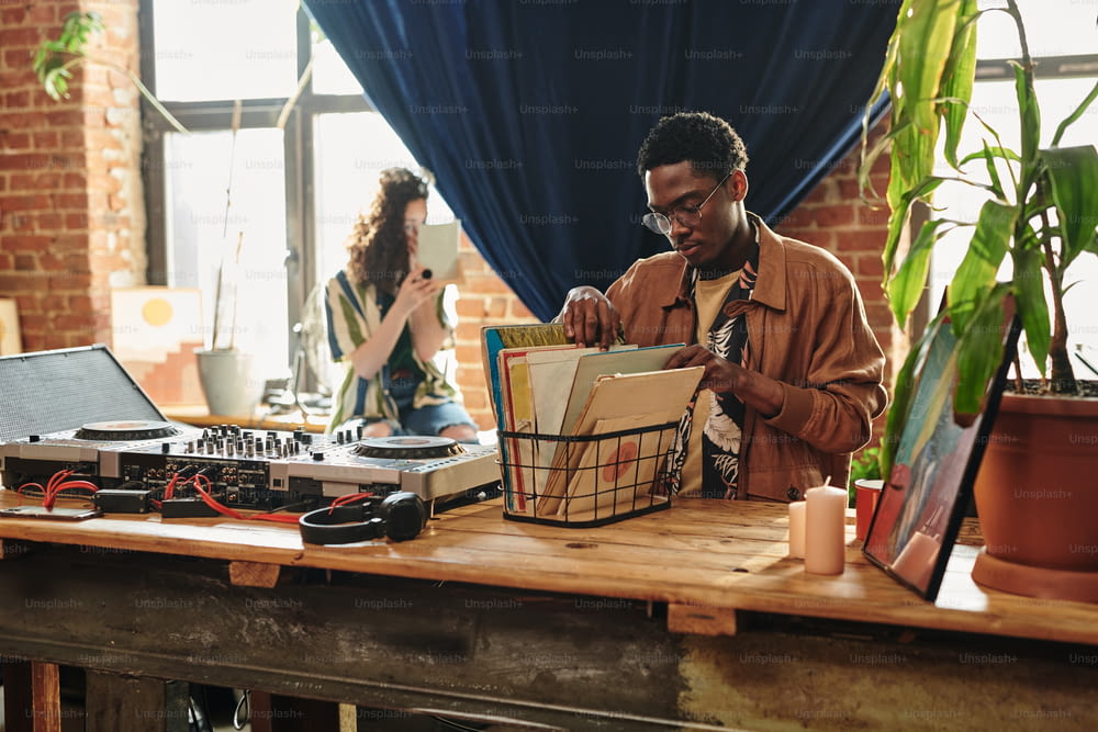 Young African American man looking through his collection of vynil disks in box standing on table next to deejay booth and headphones