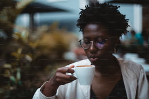 Un ritratto discreto di un'affascinante giovane donna nera con riccioli afro, in occhiali eleganti e un trench bianco, che beve una deliziosa cioccolata calda in un caffè notturno buio; uno spazio di copia posto a sinistra