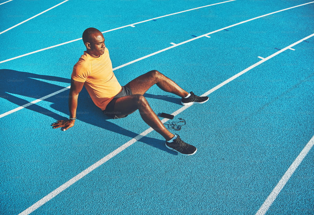 Fit young African runner sitting alone with his cellphone on a track preparing for a run on a sunny day