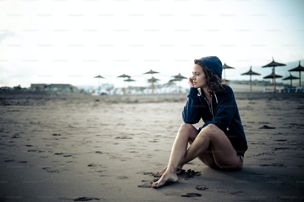 Cute lonely caucasian young girl sitting at the beach on the sand looking the open space and thinking - closed resort with sun umbrellas in background - cold tones and colors