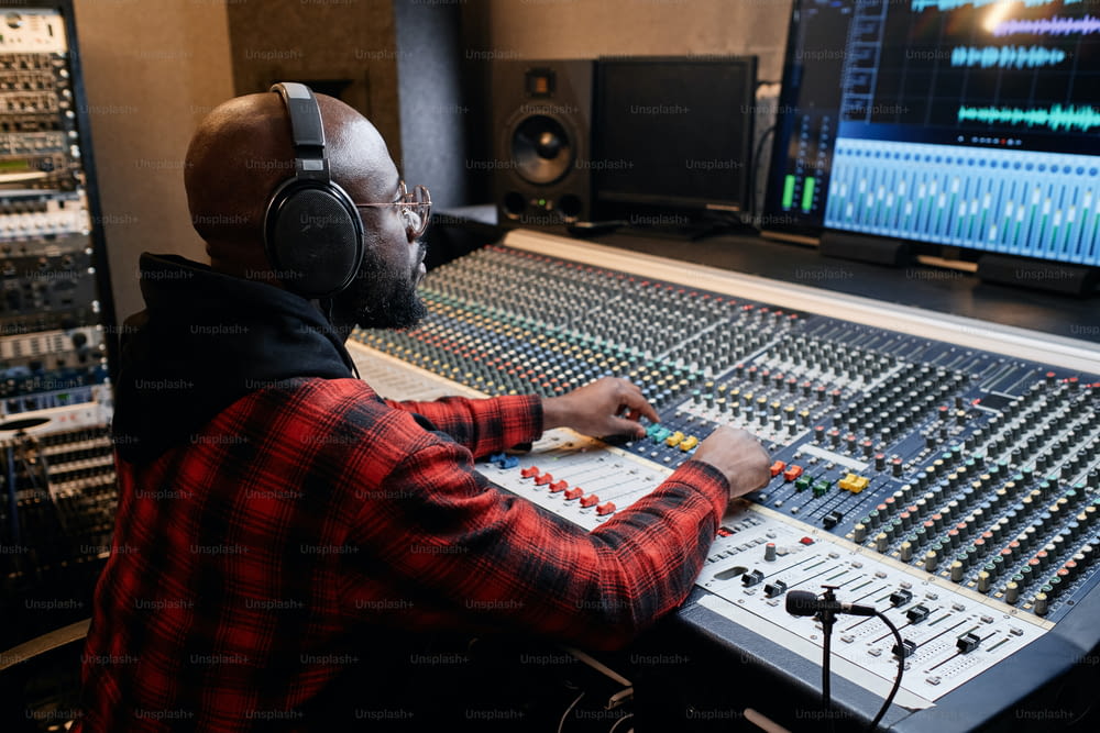 Stylish African American man wearing red checked shirt and headphones working on music track in recording studio