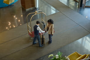 Above angle of young female traveler passing her suitcase to bellboy with cart standing in front of her in spacious lounge of hotel
