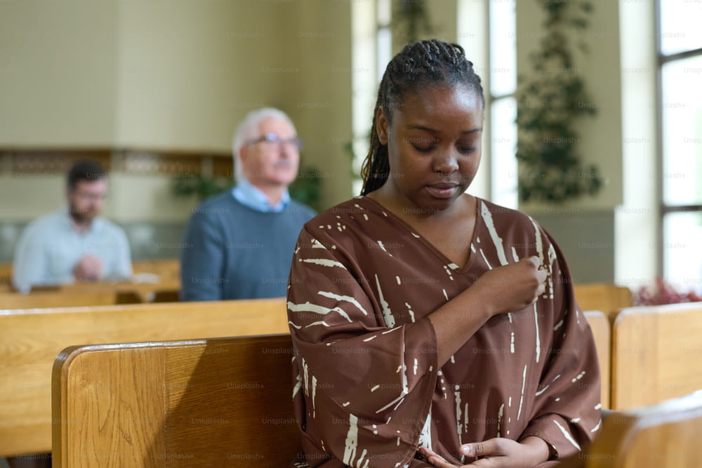 Young black woman with her eyes closed crossing herself during silent prayer while sitting on bench against parishioners in church