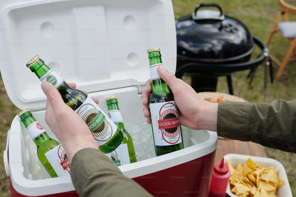 Mãos de jovem segurando duas garrafas de cerveja sobre caixa preparada para convidados e festa de quintal contra churrasqueira e batatas fritas em tigela