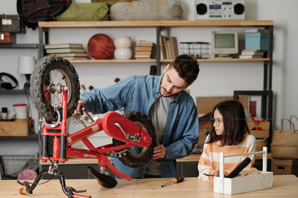 Young contemporary man checking wheels of his daughter bicycle in garage while both standing by wooden table with handtools