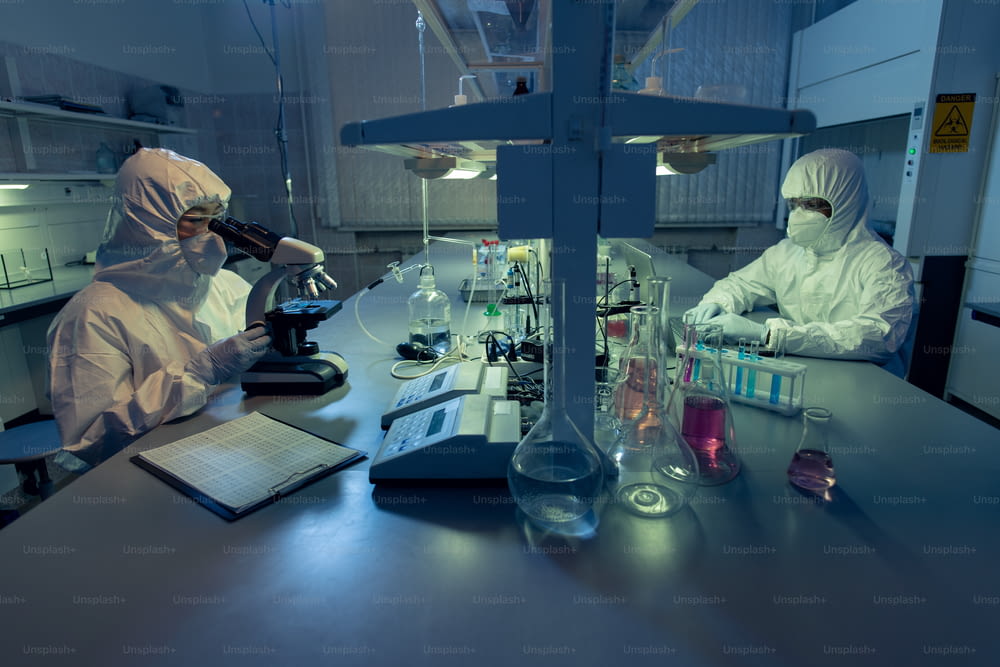 Deux collègues en tenue de travail de protection assis à la table et travaillant avec des échantillons chimiques dans des tubes à essai dans le laboratoire