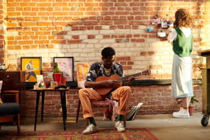 肘掛け椅子に座ってアコースティックギターを弾く若い男性と、レンガの壁の前に立って写真を見ているガールフレンド