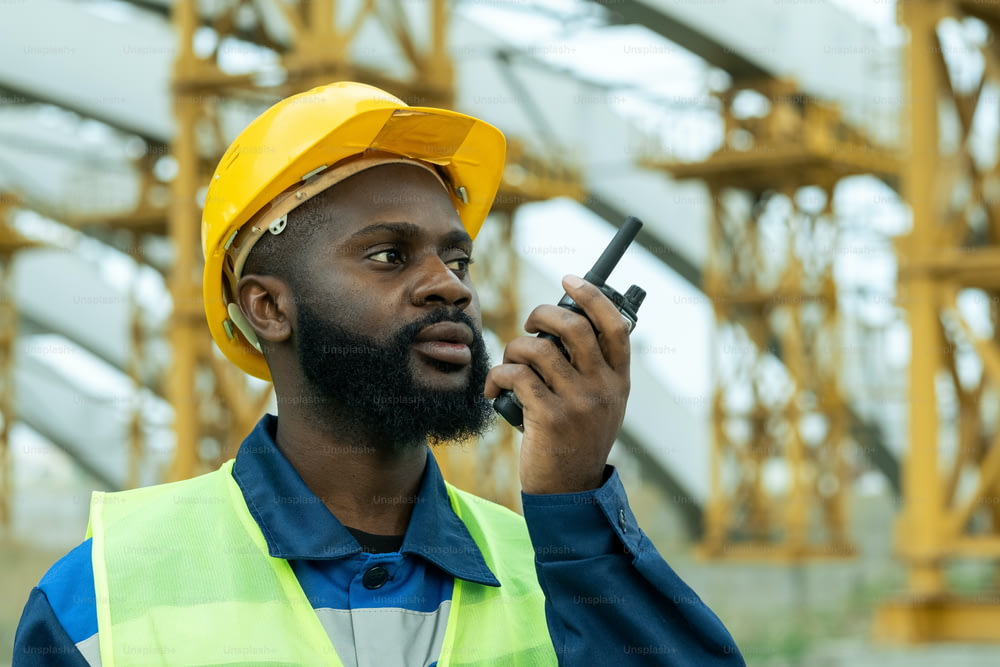 Trabalhador da construção civil africano no capacete de trabalho falando no transmissor enquanto trabalha no canteiro de obras ao ar livre