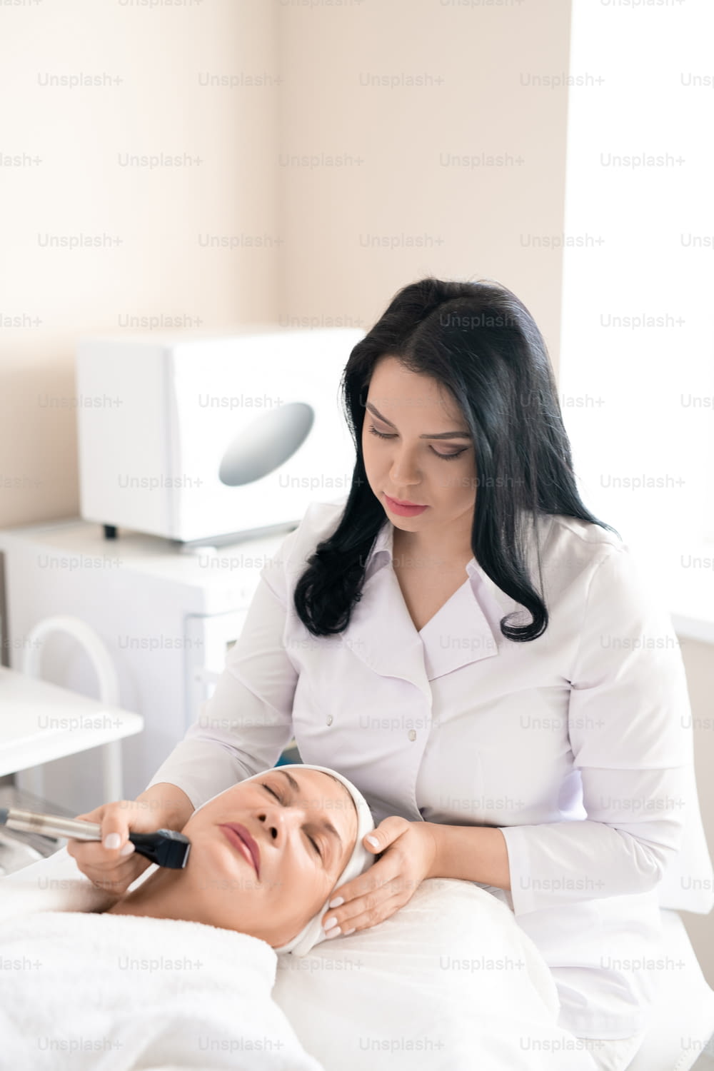 Concentrated cosmetologist in white coat using facial machine while doing skin tightening procedure to mature woman
