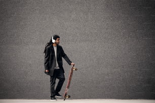 Stylish young dreadlocks hipster skater with headphones holding skateboard near the grey wall in a suit.
