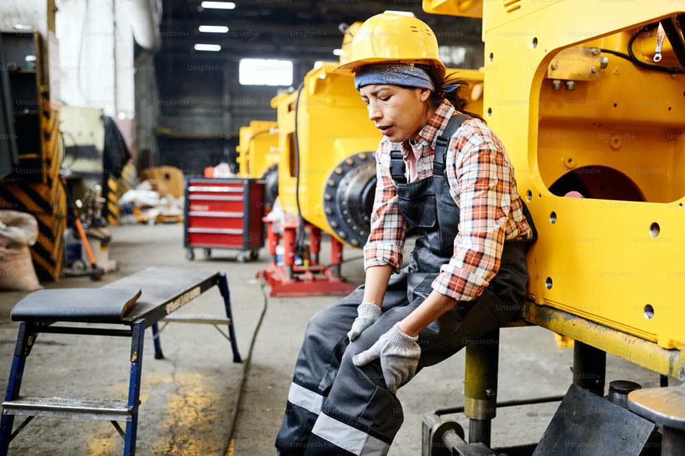Jeune ouvrière d’un entrepôt en combinaison et casque de sécurité touchant un genou blessé alors qu’elle était assise près d’une machine industrielle