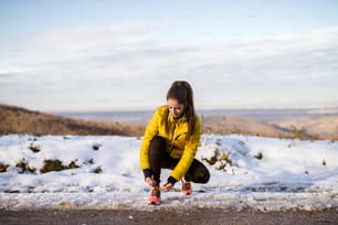 Joven delgada atlética atando cordones de zapatos en ropa deportiva de invierno en carretera invernal nevada con auriculares en la mañana soleada.