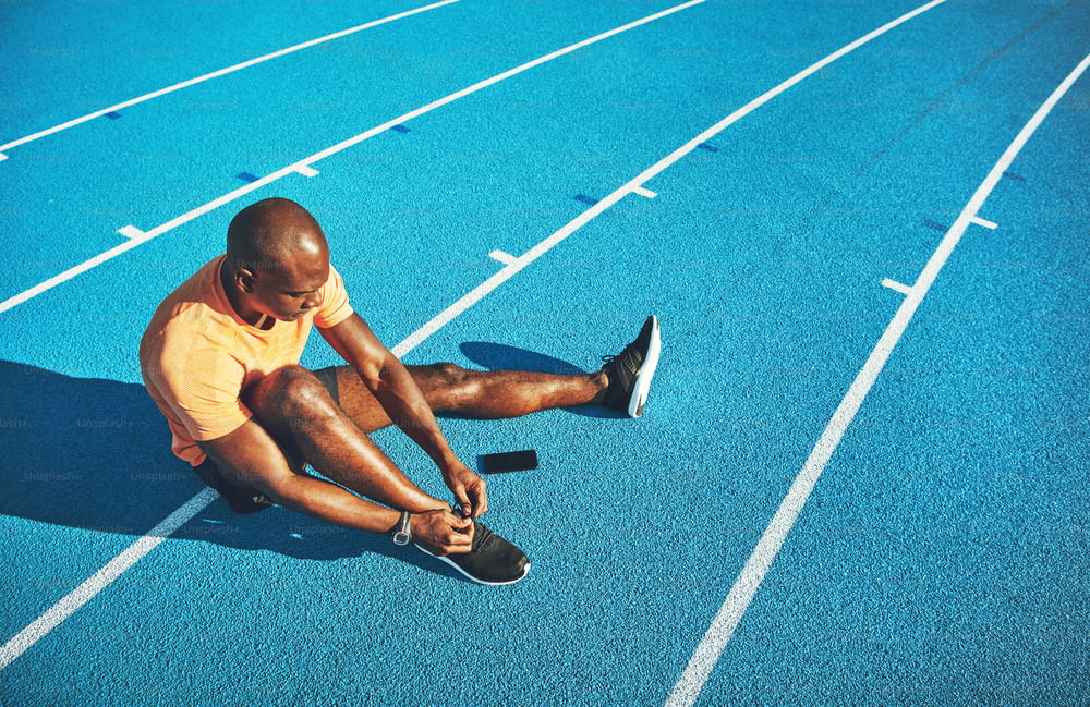 Focused young African male athlete sitting alone on a running track tying up his shoes before training